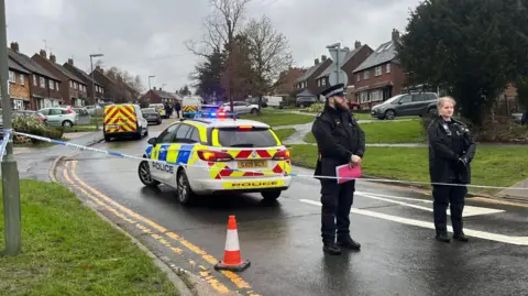 Two police officers stood behind a cordon in a road. A police car is behind them and houses line the street