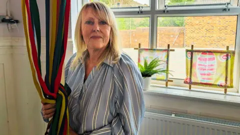Alex Moss/BBC A smiling woman holding colourful ribbons hanging from a pole. She has blonde hair and is wearing a blue and white striped shirt. She is stood in front of a window with colourful drawings on display. 