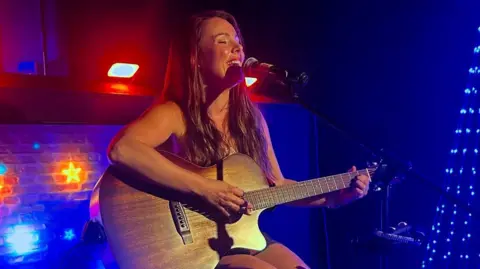 A woman with long brown hair, is sitting down on a stool on a stage while singing into a microphone and holding a guitar.