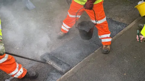A close up view of workmen in bright orange overalls holding a bucket of tar and a tool to fix a pothole on the road