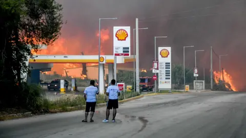 Reuters Two men stand in the road watching a wildfire billow smoke behind a petrol station.