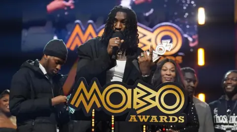PA Media Odeal wins the Best R&B/Soul Act Award at the Mobo Awards at the Utilita Arena Newcastle. He holds a mic in one hand and his trophy in the other while speaking to the crowd after the win. He has shoulder-length braided hair and wears a black blazer over a white vest. He's pictured behind a podium reading 'Mobo Awards' with some supporters behind him.