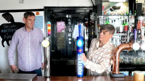 Sam Philpott está detrás del bar con su esposa Suzanne. Sam lleva una camisa a cuadros. Suzanne, que lleva una camisa estampada crema y marrón, tiene su brazo descansando sobre un toque de cerveza fomenta el bar en el bar