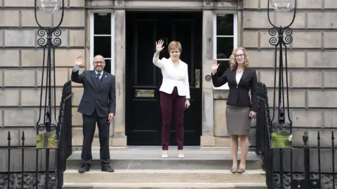 PA Media Nicola Sturgeon is flaked by Patrick Harvi and Lorna Slater on the steps at Bute House, as the three politicians wave to the camera.