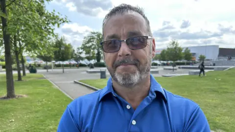Jason Lucker in a park. There is a public square behind him as well as a large Sainsbury's store. He is wearing dark sunglasses and a blue polo shirt.
