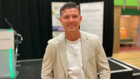Luke wears a relaxed stone coloured jacket over a white t-shirt and is smiling for the camera. A black stage is in the background.