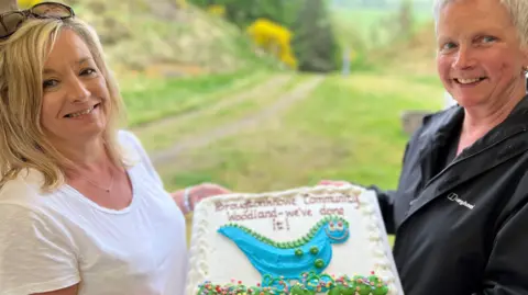 A woman with blonde hair, wearing a white T-shirt, and a grey-haired woman with a black jacket, hold a large cake with a woodland track behind them
