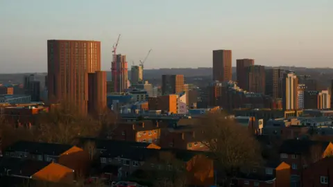 A skyline picture of Leeds city centre. The picture is taken at dawn, with the sun illuminating apartment buildings.