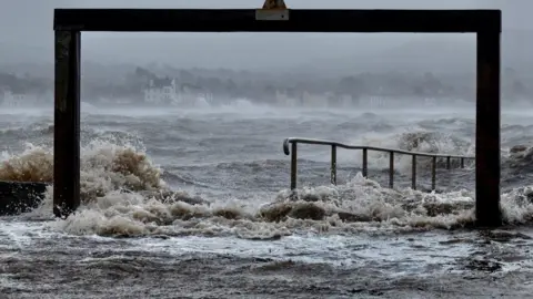Ann Bruen Large waves crash around pillars and steps on the seafront. A village is in the background on the far shore
