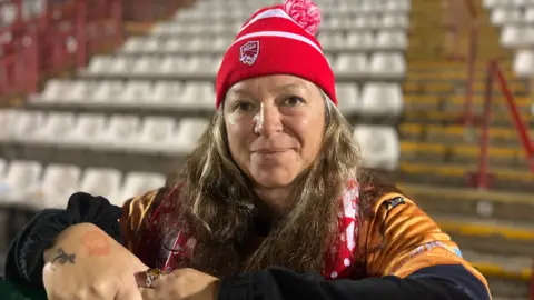 Sally looks at the camera as she wears a red Jersey Bulls beanie hat and scarf. She has a yellow football top on with a black jumper under it. She has long light brown hair which comes past her shoulders.