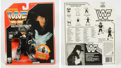 Vectis Auctions The Undertaker toy in its packaging. The figure is dressed all in black and is wearing a large black hat. The car is red with a picture of the Undertaker. The back of the card shows other figures available.