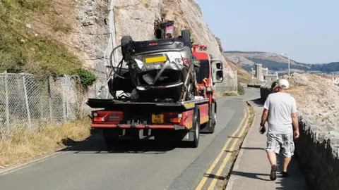 FPNW Upturned silver car fixed to back of red low loader and driven away as passersby look on