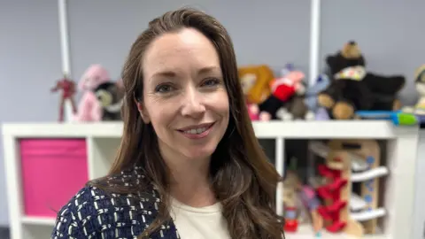 A woman with brown long hair smiles at the camera, she is wearing a white top and a blue and white jacket, and is standing in front of a unit filled with children's toys.