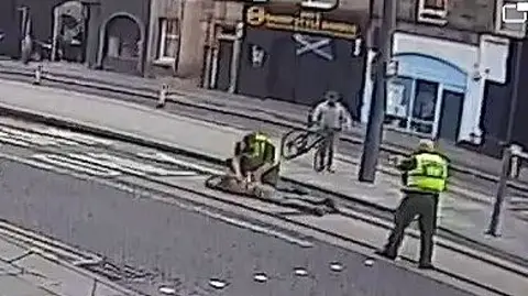 BBC A man is lying on the ground being restrained by police officers in a street