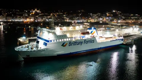 A large white and blue Brittany Ferries ship is docked in a harbour at night, brightly lit against dark water. City lights from buildings on the hillside glow in the background, reflecting across the harbour, while the ferry’s ramps and terminal structures extend alongside the vessel.