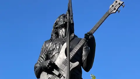 A black-coloured statue of Lemmy holding a bass guitar and singing up towards a microphone