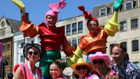 Seven people smiling and looking at the camera at a Pride event. Two people are dressed in colourful costumes, holding their arms out, and three people are wearing hats. A building is behind them.