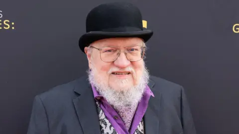 Getty Images George RR Martin with grey beard, glasses, bowler hat, purple shirt, patterned waistcoat and grey suit jacket, looking at the camera and smiling.