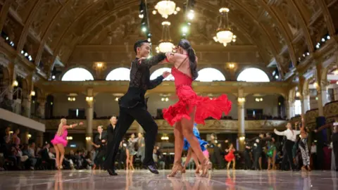 Getty Images A ballroom dancing couple wearing black and red dancing in the centre of a large dancefloor.