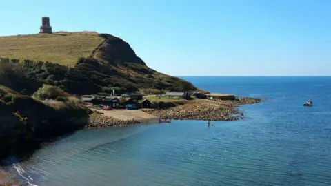 Oast House Archive / Geograph View across Kimmeridge Bay. To the left of the picture is the cliff with Clavell Tower - a round folly - at the top. Beneath is a slipway with some buildings at the top. In the foreground is the shore. The right of the photo is just clear blue sky and vivid blue sea.