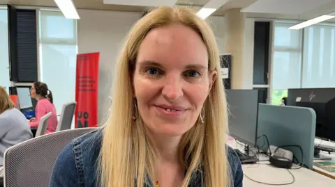 Ollie Conopo/BBC A woman with long blonde hair looks at the camera and smiles. She is wearing a denim jacket.