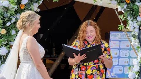 Rachel Clark Rachel Clark reading from a black book with a woman standing to her left in a wedding dress 