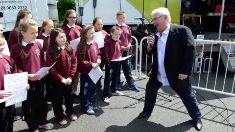 Eleven primary school students on the left alongside Hugo Duncan on the right. He is speaking into a microphone as he broadcasts live from St Peter's School as as they celebrate the school's centenary as part of BBC Music Day. The pupils are wearing a burgundy sweater, white shirt and black trousers with trainers. Mr Duncan is wearing a white shirt, black blazer and black shoes.