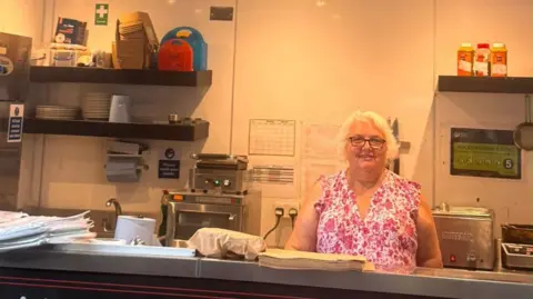 Maria's Market Cafe Maria Moruzzi looks into the camera and smiles. She is visible from the waist up and is wearing a white and pink patterned dress or blouse. She has white hair and dark-framed glasses. She is standing behind a cafe counter with various items of catering equipment on shelves and a counter behind her.