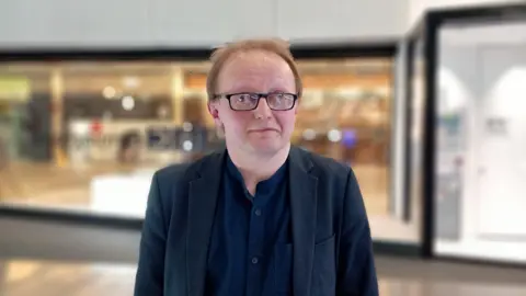 A man with blonde/grey hair stands outside a glass facade in a shopping centre. He is wearing a dark blue suit, dark blue shirt and black-rimmed glasses. The background is blurred but is brightly lit. 