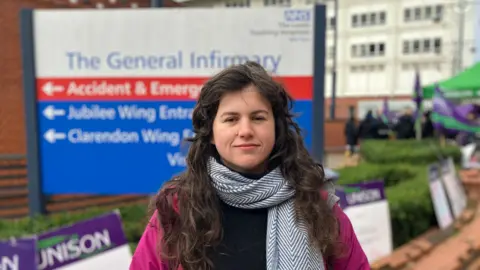 A woman standing in front of The Leeds General Infirmary sign, dressed warmly in a magenta jacket and a grey and white scarf.