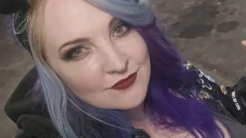 Jaz Morse Jaz smiles at the camera in a selfie. She is wearing a black hoodie with lots of badges on it, and has multi-coloured purple hair.