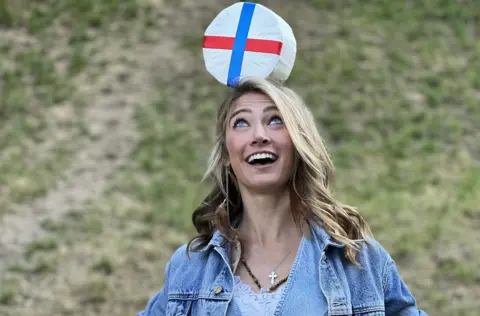 A woman with blonde hair and wearing a blue denim jacket smiles as she balances a round double gloucester cheese on her head