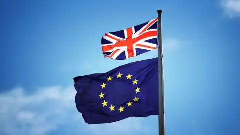 A small red, white and blue British Union Jack blows in the breeze above a a European Union flag, which consists of a ring of yellow stars against a block blue background. The sky in the background is clear. 