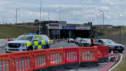 Helen Mulroy/BBC A police truck has its blue lights on behind a line of orange barriers shutting the road. It is blocking the entrance to a tunnel at the airport, which has a stream of cars queuing to get out.