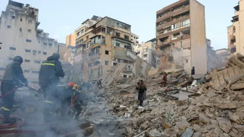REUTERS/Mohamed Azakir Emergency responders in uniforms and helmets hold a hose as they stand in the smoking rubble of a collapsed building. Other buildings remain standing nearby.