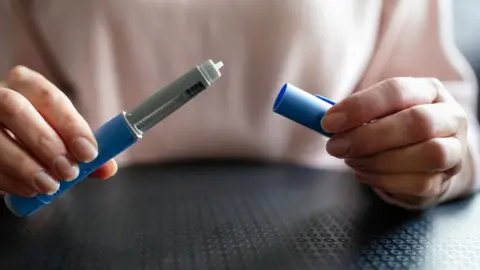 The Getty Images stock photo shows a close-up of a man holding two pieces of a weight loss jab, the jab looks like a pen with the cap removed, and the man is wearing a pink top and sitting at a table inside.
