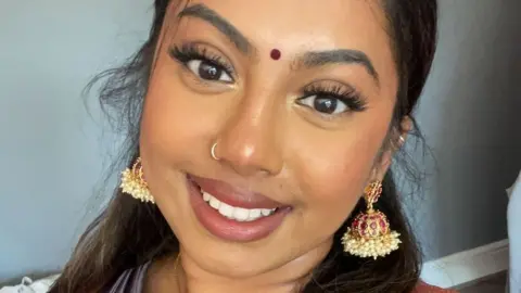 Kirthana  Kirthana - a 21-year-old south Asian woman, smiles at the camera as she takes a selfie. She has her long, dark hair styled half-up-half-down and wears a bindi and her makeup includes a glittery gold eyeshadow and false eyelashes. She wears decorative dangly earrings which comprise a round red ornament decorated in gold and pearls. She is pictured inside against a pale blue wall.