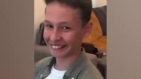 The family smiles while watching a young boy camera. He appears in a living room. He is wearing a khaki shirt over a white T-shirt.