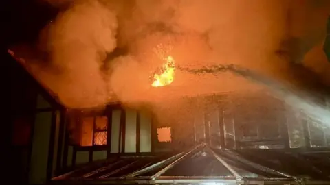 Essex Fire Service A jet of water attempts to douse flames on a large derelict house which is on fire.