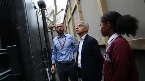 PA Media The mayor is stood looking up at a large black heat pump, alongside the headteacher who is wearing a blue shirt, a school pass and glasses and a female pupil wearing a dark red cardigan is also stood by them. 