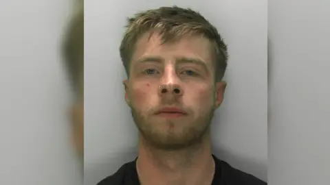 Gloucestershire Police Mugshot of Stack who has blonde hair, a nose ring, and blue eyes. He is wearing a black top.