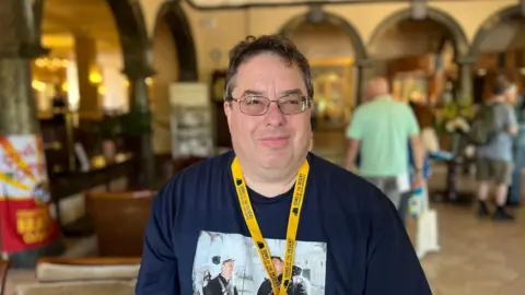 A man wearing a blue T-shirt and a yellow lanyard looks to the right of the camera. He is wearing glasses.
