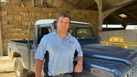 Jack Harvey, a man with short, dark hair and a light blue polo shirt, is standing in front of a pick-up truck and a wall of hay bales.