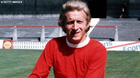 Dennis Law in a long sleeved red Manchester United shirt with white round neck collar.  Football pitch and terrace visible behind.