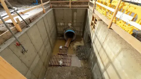 Aerial shot of the pipe. The pipe is deep below the ground. Wooden slats are on the floor in front of the entrance to the pipe. 