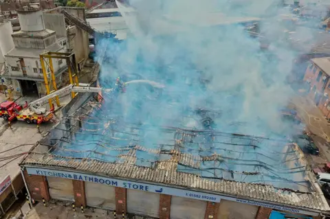 Stephen Huntley/BBC Two firefighters on an aerial platform spraying water over the what is left of the roof of a building. Only the blackened frame of the roof remains and smoke is rising from it. There is an industrial building to the left. 