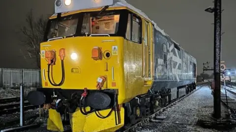 Nigel Hanson A locomotive in the dark with a yellow front and dark blue rear with a small yellow snow plough attached to the front and snow on the ground all around the tracks
