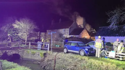 NFRS Night time image of fire in a house in a village. Smoke and flames can be seen in the background. Fire crews in the foreground. There are barn-type structures in view and a small bridge running over a stream.