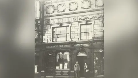 An undated black and white image of the exterior of the Angel Inn in Stockport with a man and a woman standing at the entrance.