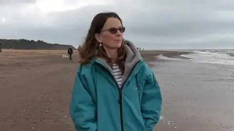 A woman wearing a long blue jacket walking along the beach with the sea to her right.